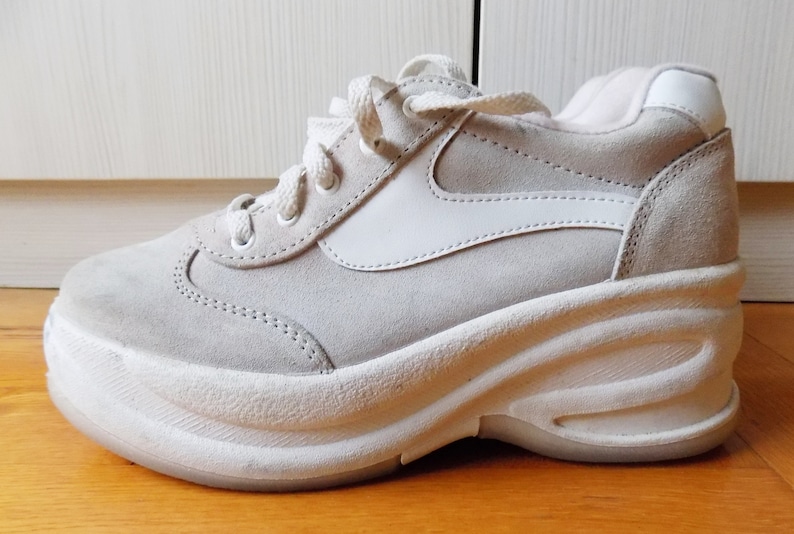 May include: A light gray platform sneaker with white accents. The shoe features a suede upper, a thick, wavy white sole, and white laces. The design includes a white side panel and a padded collar.