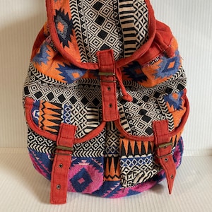 Może przedstawiać: Kolorowy plecak z geometrycznym wzorem w kolorze pomarańczowym, niebieskim, czarnym i białym. Plecak ma dwie przednie kieszenie oraz czerwone paski i akcenty. Torba ma zapięcie na sznurek i regulowane paski.