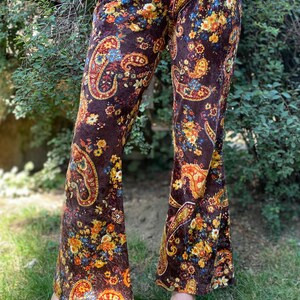 May include: Brown velvet bell-bottom pants with a paisley and floral pattern in shades of orange, yellow, and blue. The pants have a high waist and are worn with black platform sandals. The pants are made of a soft, flowing fabric.