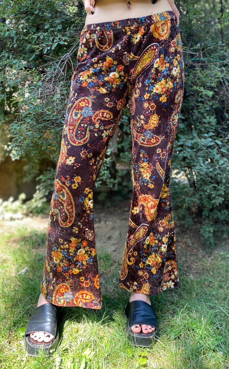 May include: Brown velvet bell-bottom pants with a paisley and floral pattern in shades of orange, yellow, and blue. The pants have a high waist and are worn with black platform sandals. The pants are made of a soft, flowing fabric.