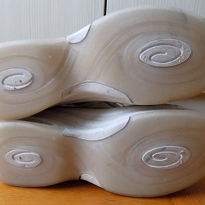 May include: Close-up of the soles of two athletic shoes. The soles are a translucent off-white color with a swirl design and a circular logo. The shoes are resting on a wooden surface.