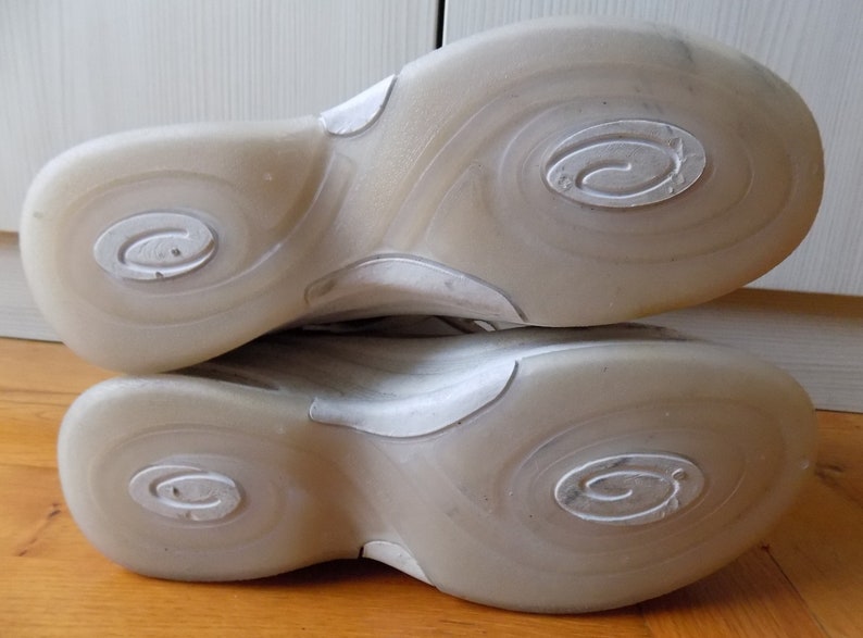 May include: Close-up of the soles of two athletic shoes. The soles are a translucent off-white color with a swirl design and a circular logo. The shoes are resting on a wooden surface.
