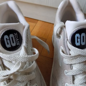 May include: Pair of white sneakers with white laces. Each shoe features a black circular emblem with the text "GO RIVOX" in light blue. The sneakers are set against a light brown wooden surface.