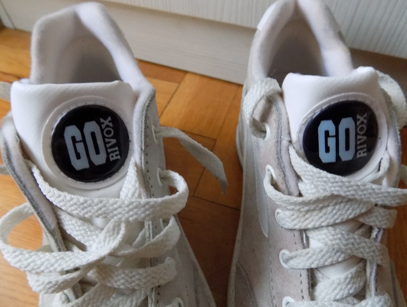 May include: Pair of white sneakers with white laces. Each shoe features a black circular emblem with the text "GO RIVOX" in light blue. The sneakers are set against a light brown wooden surface.