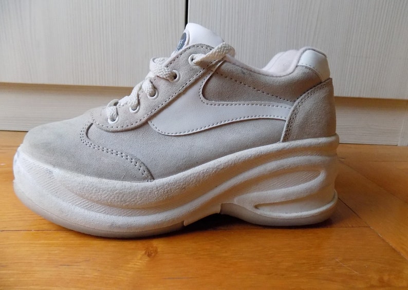 May include: A light gray suede sneaker with a thick white sole. The shoe features a white accent on the side and white laces. The sneaker has a retro style and is suitable for casual wear.