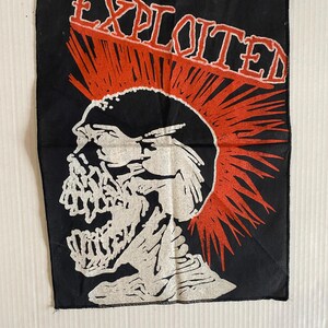 Pode incluir: Patch preto com um desenho de caveira branca e um moicano vermelho, com a palavra "EXPLOITED" em vermelho. O patch é retangular e feito de tecido, projetado para ser costurado ou passado a ferro em roupas ou acessórios.