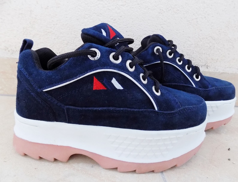 May include: Navy blue platform sneakers with white and red accents. The shoes feature a suede upper, white soles, and pink rubber outsoles. The sneakers have black laces and silver eyelets. A stylish and comfortable footwear option.