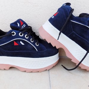 May include: Navy blue platform sneakers with white and pink accents. The shoes feature a suede-like texture, white laces, and a logo with red and white details. The thick sole is white with a pink bottom.