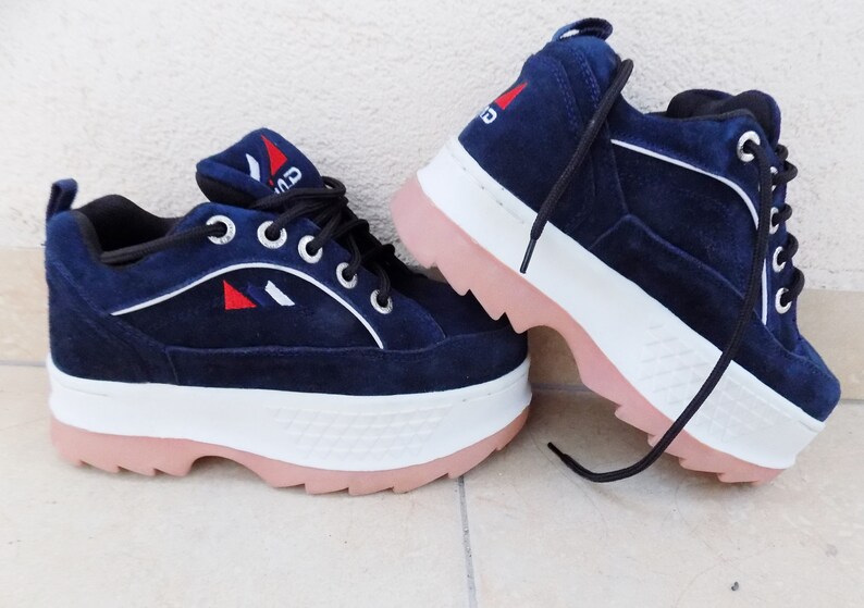 May include: Navy blue platform sneakers with white and pink accents. The shoes feature a suede-like texture, white laces, and a logo with red and white details. The thick sole is white with a pink bottom.