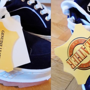 May include: Navy blue sneakers with white laces and soles. The shoes have a pink sole trim and a tag that reads "GENUINE LEATHER". The tag also lists features like "durable soft leather upper" and "long-lasting comfort".