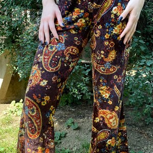 May include: Brown paisley print pants with a flared leg. The pants feature a repeating pattern of large paisley shapes in shades of gold, blue, and orange. The fabric appears to be velvet or a similar soft material.