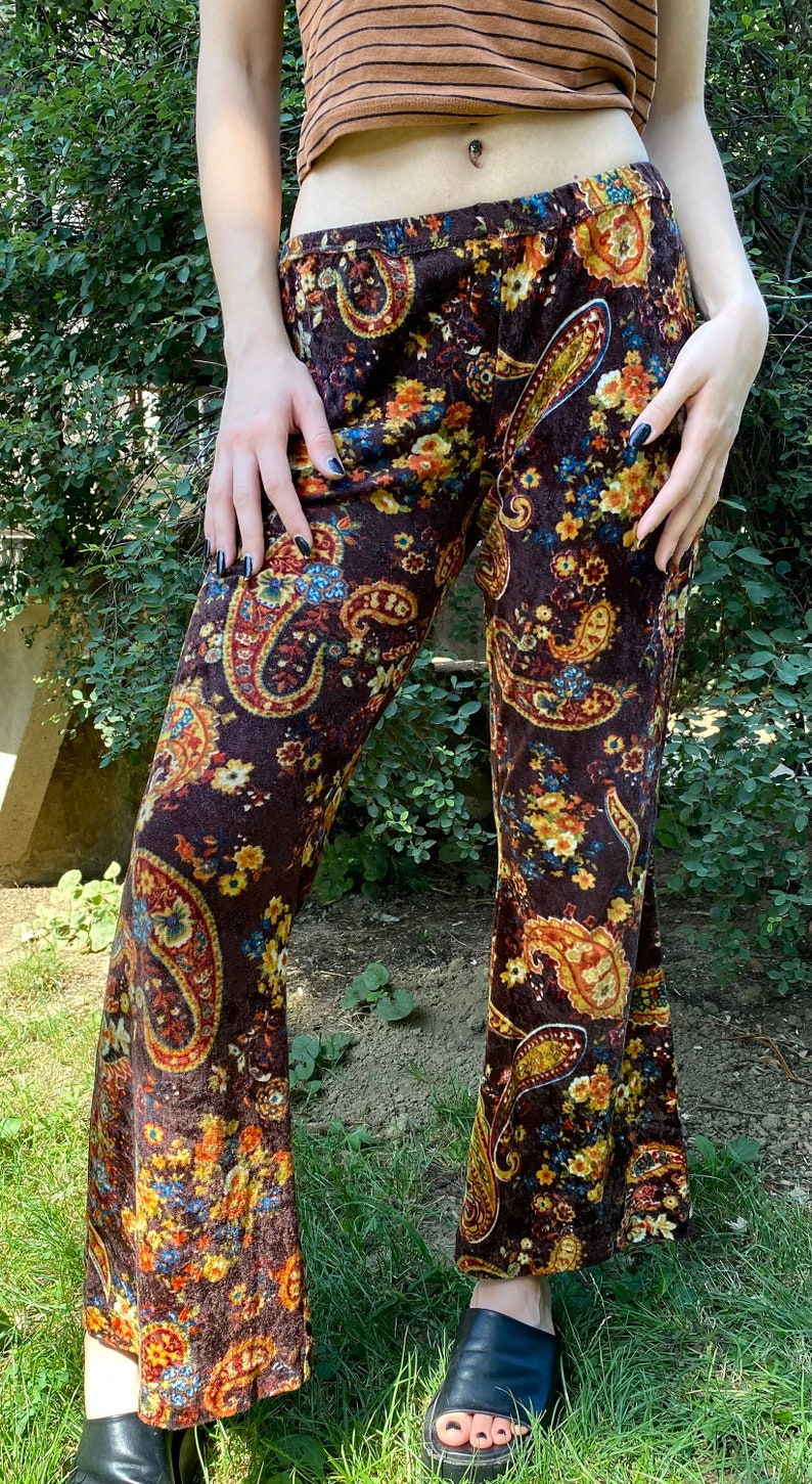 May include: Brown paisley print pants with a flared leg. The pants feature a repeating pattern of large paisley shapes in shades of gold, blue, and orange. The fabric appears to be velvet or a similar soft material.