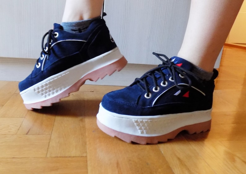 May include: Navy blue platform sneakers with white and pink accents. The shoes feature a suede-like upper, white soles with a geometric design, and pink rubber outsoles. The sneakers have black laces and silver eyelets.