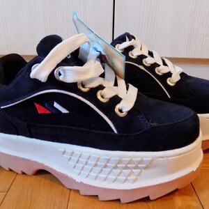 May include: Navy blue platform sneakers with white laces and a white and tan sole. The sneakers have gold eyelets and a small logo with red, white, and blue accents. The shoes have a suede-like texture.