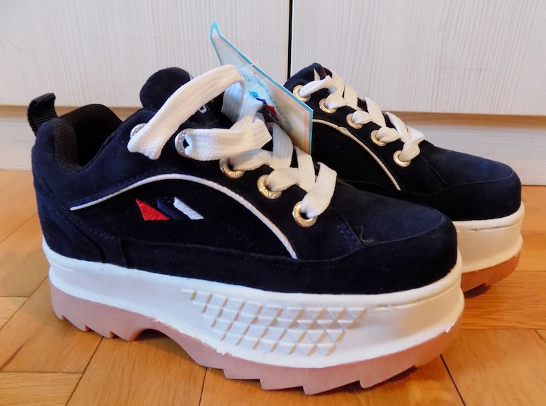 May include: Navy blue platform sneakers with white laces and a white and tan sole. The sneakers have gold eyelets and a small logo with red, white, and blue accents. The shoes have a suede-like texture.