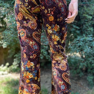 May include: Brown velvet flared pants with a colorful floral and paisley pattern. The pants feature a high waist and a wide leg, with a vibrant mix of orange, yellow, and blue floral designs. The fabric has a soft, textured appearance.
