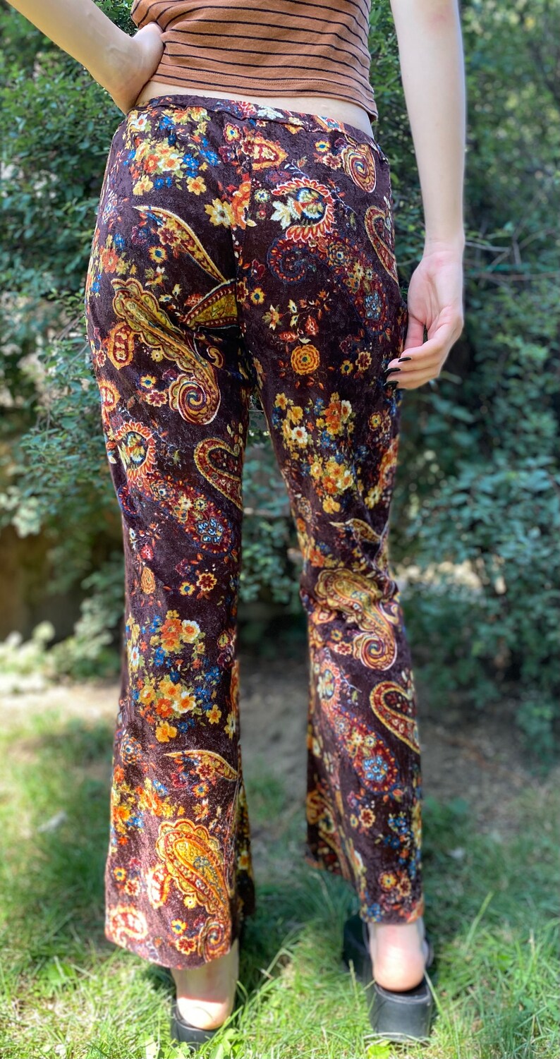 May include: Brown velvet flared pants with a colorful floral and paisley pattern. The pants feature a high waist and a wide leg, with a vibrant mix of orange, yellow, and blue floral designs. The fabric has a soft, textured appearance.