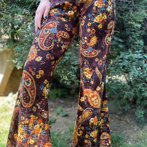 May include: Brown velvet bell-bottom pants with a paisley and floral pattern in shades of yellow, orange, and blue. The pants have a high waist and a wide flare from the knee down. The fabric appears soft and has a slight sheen.