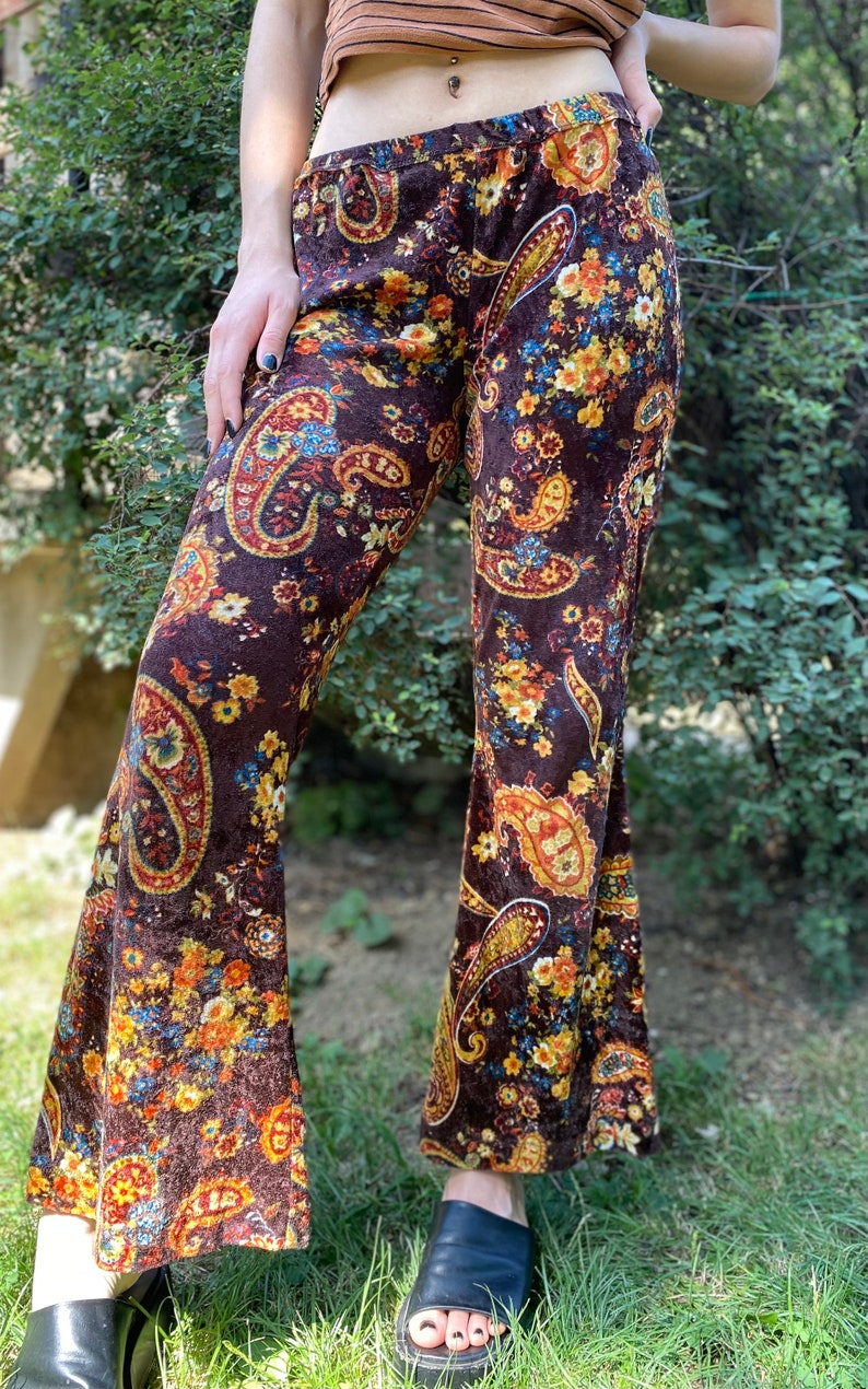 May include: Brown velvet bell-bottom pants with a paisley and floral pattern in shades of yellow, orange, and blue. The pants have a high waist and a wide flare from the knee down. The fabric appears soft and has a slight sheen.