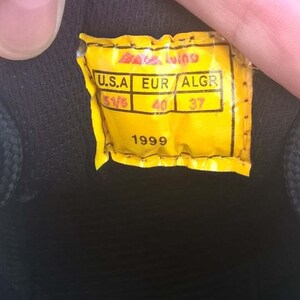 May include: Close-up of a yellow tag with black text and a black background. The tag reads "U.S.A 5 1/5, EUR 40, ALGR 37, 1999". The tag is attached to a black shoe.