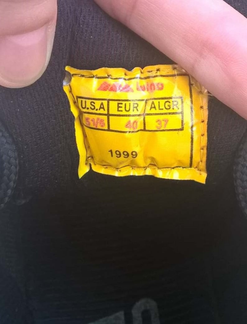 May include: Close-up of a yellow tag with black text and a black background. The tag reads "U.S.A 5 1/5, EUR 40, ALGR 37, 1999". The tag is attached to a black shoe.