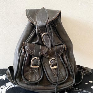 Op de afbeelding: Een donkerbruine leren rugzak met twee voorzakken, zilveren gespen en witte stiksels. De rugzak heeft een trekkoordsluiting en een klep met decoratieve stiksels. De tas staat op een zwart-wit patroon oppervlak.