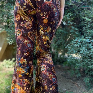 May include: Brown velvet bell-bottom pants with a colorful paisley floral pattern. The pants feature a high waist and a flared leg, with a mix of orange, yellow, and blue floral designs.