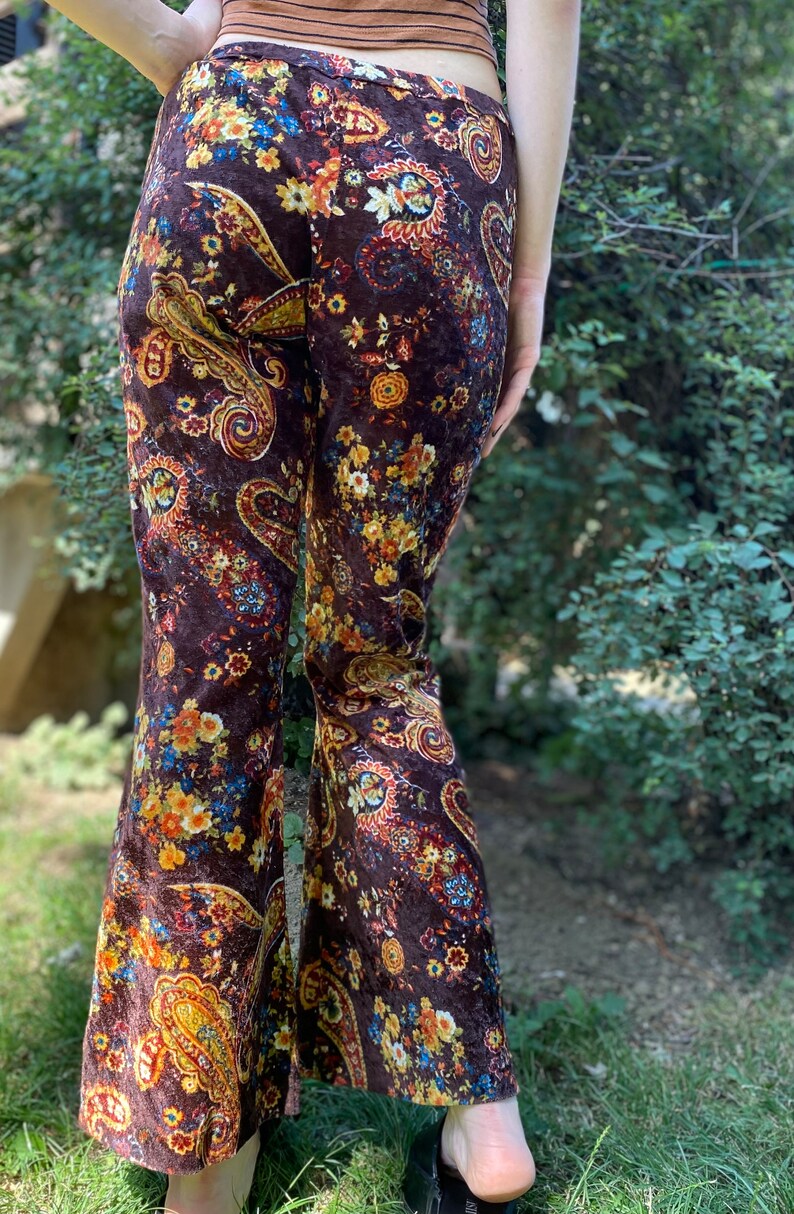 May include: Brown velvet bell-bottom pants with a colorful paisley floral pattern. The pants feature a high waist and a flared leg, with a mix of orange, yellow, and blue floral designs.
