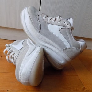 May include: A pair of light gray sneakers with white accents. The shoes have a thick, white sole and a suede-like upper. The design includes white side panels and laces. The sneakers are shown on a wooden floor.