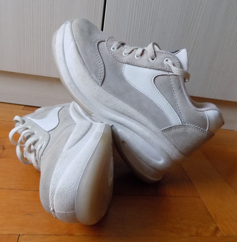 May include: A pair of light gray sneakers with white accents. The shoes have a thick, white sole and a suede-like upper. The design includes white side panels and laces. The sneakers are shown on a wooden floor.