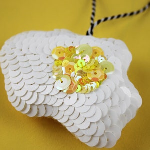May include: A white cloud-shaped ornament with a yellow and white sequin design. The ornament is hanging from a black and white string.