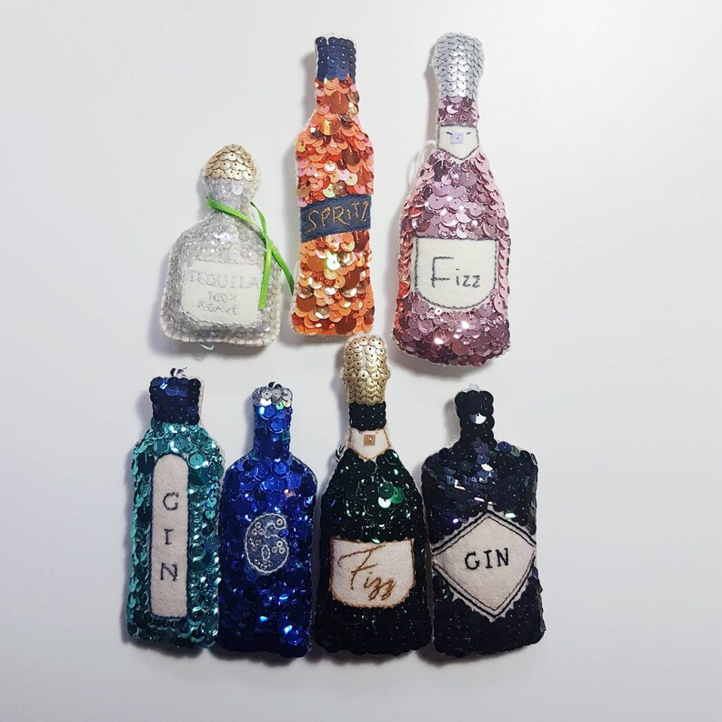 K&ouml;nnte beinhalten: Sieben Miniaturflaschen, verziert mit Pailletten und Stickerei. Die Flaschen sind mit "Gin", "Fizz", "Spritz", "Fizz" und "Gin" beschriftet.