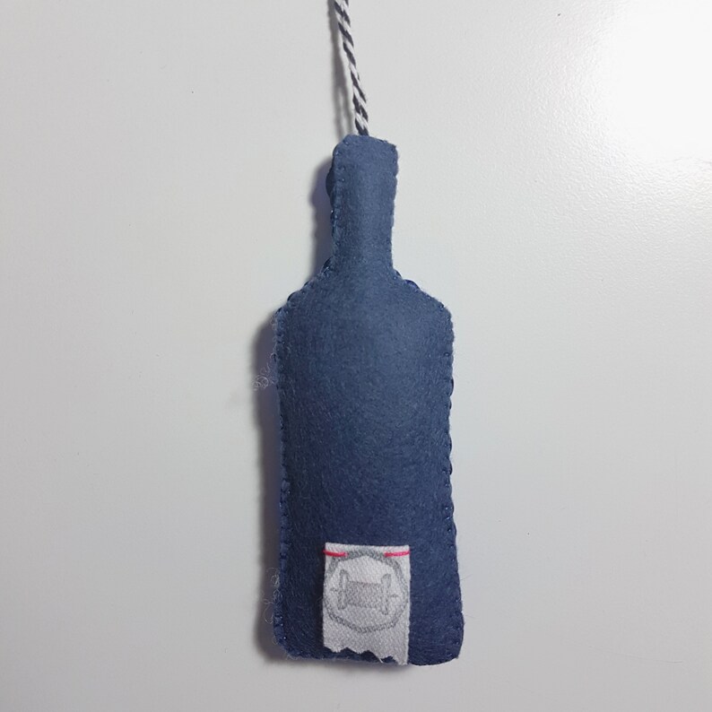 K&ouml;nnte beinhalten: Ein blauer Filzflaschen-Anh&auml;nger mit einem wei&szlig;en Etikett und einem gestickten Design. Der Anh&auml;nger h&auml;ngt an einer wei&szlig;en und schwarzen Schnur.
