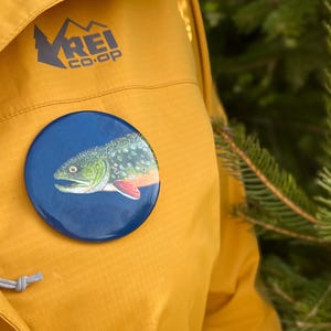 May include: A yellow jacket with the REI co-op logo on the left chest. A round pin with a blue background and a brown trout with red spots is pinned to the jacket.