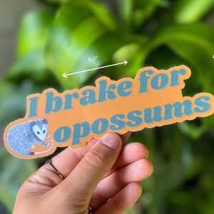 Op de afbeelding: Een gestanste sticker met een illustratie van een grijze opossum en de blauwe tekst "I brake for opossums" op een oranje achtergrond. De sticker is 16,51 cm breed en 5,13 cm hoog, zoals aangegeven door de zichtbare maatpijlen op de afbeelding.
