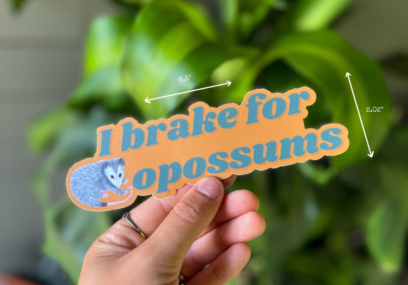 Op de afbeelding: Een gestanste sticker met een illustratie van een grijze opossum en de blauwe tekst "I brake for opossums" op een oranje achtergrond. De sticker is 16,51 cm breed en 5,13 cm hoog, zoals aangegeven door de zichtbare maatpijlen op de afbeelding.