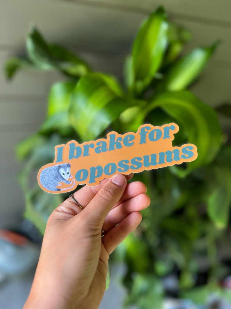 Op de afbeelding: Een rechthoekige sticker met een oranje achtergrond en blauwe tekst die I brake for opossums leest. De sticker bevat een kleine illustratie van een grijs en wit opossum aan de linkerkant, geplaatst naast de tekst in een vetgedrukt lettertype.