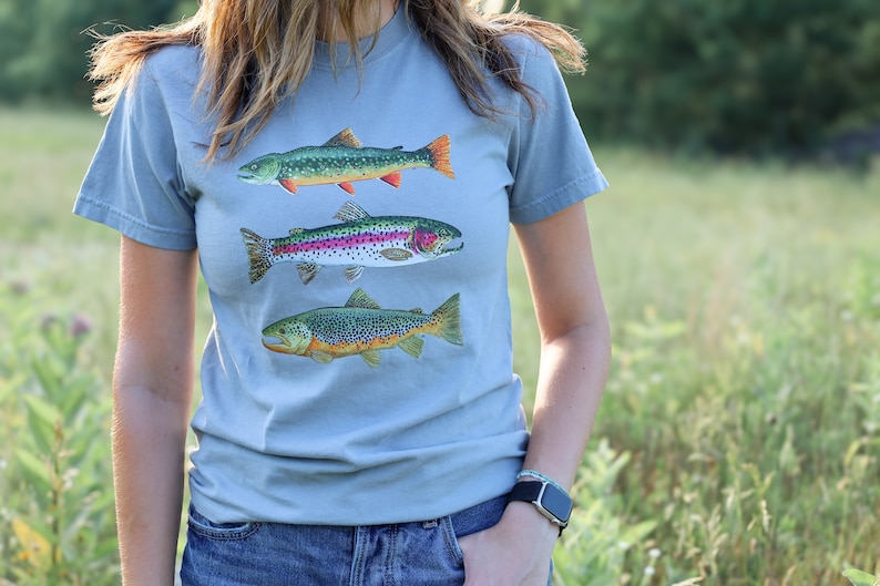 Puede incluir: Una camiseta azul claro con un gr&aacute;fico de tres tipos diferentes de truchas. Los peces est&aacute;n ilustrados en un estilo realista con marcas y colores detallados.