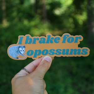 Op de afbeelding: Een gestanste sticker met een mosterdgele achtergrond met een grijze illustratie van een opossum aan de linkerkant en de blauwe tekst "I brake for opossums" in een vet, afgerond, schreefloos lettertype in het midden.