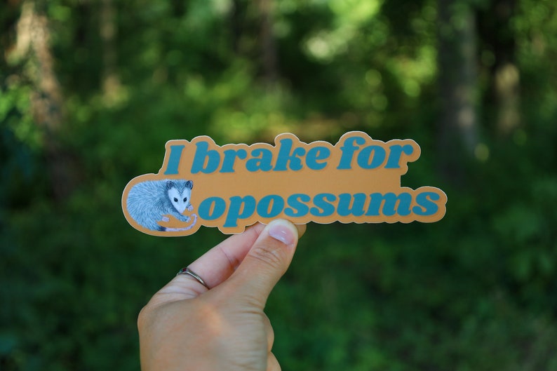 Op de afbeelding: Een gestanste sticker met een mosterdgele achtergrond met een grijze illustratie van een opossum aan de linkerkant en de blauwe tekst "I brake for opossums" in een vet, afgerond, schreefloos lettertype in het midden.