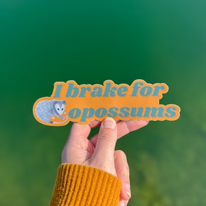 Op de afbeelding: Een gestanste vinylsticker met een oranje achtergrond en blauwe tekst die I brake for opossums leest. De sticker bevat een gedetailleerde illustratie van een grijs en wit opossum aan de linkerkant, geplaatst naast de letter I.