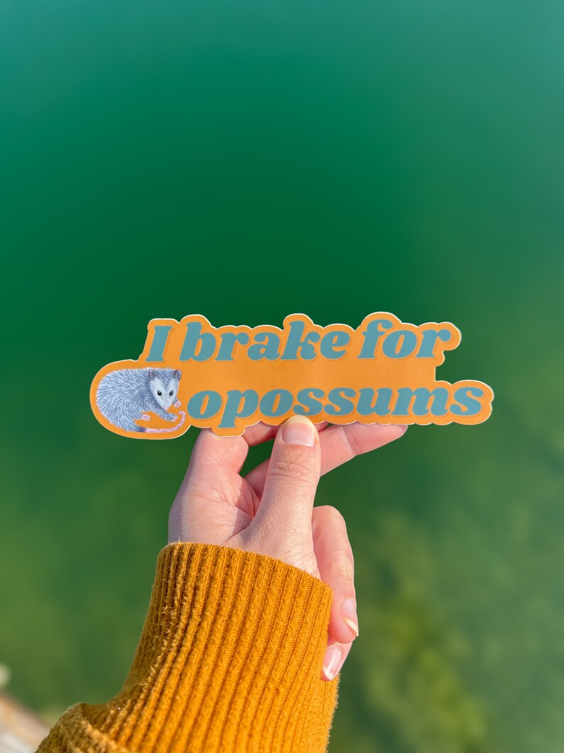 Op de afbeelding: Een gestanste vinylsticker met een oranje achtergrond en blauwe tekst die I brake for opossums leest. De sticker bevat een gedetailleerde illustratie van een grijs en wit opossum aan de linkerkant, geplaatst naast de letter I.