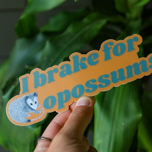 Op de afbeelding: Een gestanste sticker met een oranje achtergrond en een gedetailleerde illustratie van een grijs en wit opossum aan de linkerkant. De tekst "I brake for opossums" staat in dikke, blauwe, afgeronde letters in het midden van de sticker.