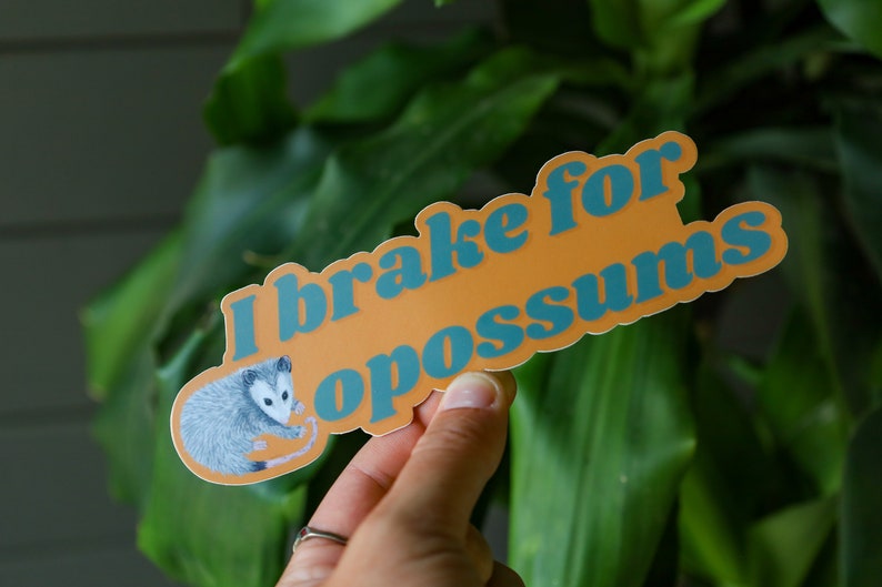 Op de afbeelding: Een gestanste sticker met een oranje achtergrond en een gedetailleerde illustratie van een grijs en wit opossum aan de linkerkant. De tekst "I brake for opossums" staat in dikke, blauwe, afgeronde letters in het midden van de sticker.