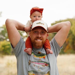 Puede incluir: Un hombre con una camiseta gris con un estampado de tres truchas. Lleva a una ni&ntilde;a peque&ntilde;a sobre sus hombros. La ni&ntilde;a lleva una diadema marr&oacute;n y una gorra de camionero marr&oacute;n y blanca con una trucha.