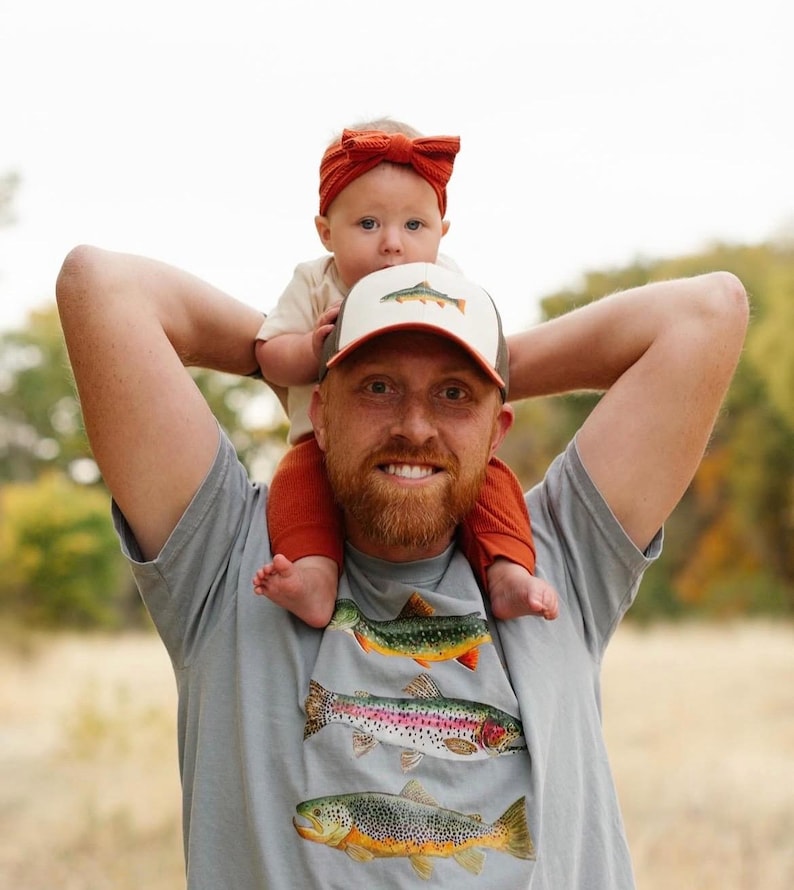 Puede incluir: Un hombre con una camiseta gris con un estampado de tres truchas. Lleva a una ni&ntilde;a peque&ntilde;a sobre sus hombros. La ni&ntilde;a lleva una diadema marr&oacute;n y una gorra de camionero marr&oacute;n y blanca con una trucha.