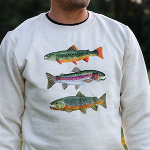 May include: A light gray sweatshirt featuring three detailed illustrations of fish. The top fish is green and orange, the middle fish is rainbow-colored, and the bottom fish is brown with orange accents. The sweatshirt is a neutral color.