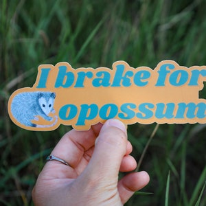Op de afbeelding: Een gestanste sticker met een mosterdgele achtergrond met een gedetailleerde illustratie van een grijs en wit opossum aan de linkerkant en de blauwe tekst "I brake for opossums" in een vet, afgerond retro-lettertype in het midden.