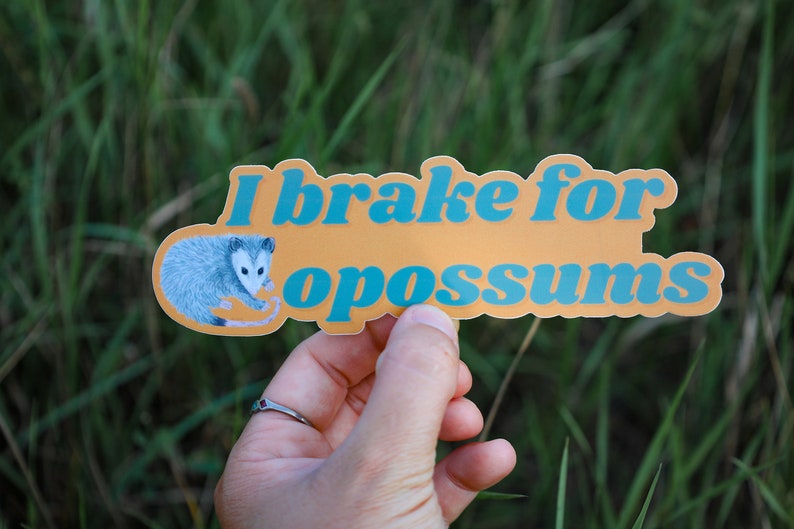 Op de afbeelding: Een gestanste sticker met een mosterdgele achtergrond met een gedetailleerde illustratie van een grijs en wit opossum aan de linkerkant en de blauwe tekst "I brake for opossums" in een vet, afgerond retro-lettertype in het midden.