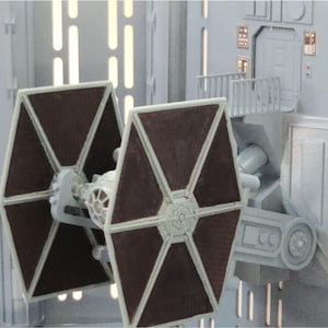 Puede incluir: Maqueta detallada de un caza TIE con paneles solares marrón oscuro y un cuerpo gris, posicionado frente a una estructura gris con una puerta y un pequeño balcón. La escena está iluminada con una luz suave y difusa.